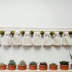 Puede incluir: Una guirnalda decorativa de borlas color crema con pompones verde oliva. Debajo, una fila de pequeñas macetas de terracota con varias suculentas. La guirnalda está fijada a una pared blanca, creando una estética minimalista y natural.