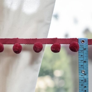 May include: A dark red pom-pom trim featuring a series of small, round, fuzzy spheres attached to a flat ribbon edge. A blue measuring tape is positioned vertically against the trim, showing a width of approximately 2 centimetres for the pom-poms.