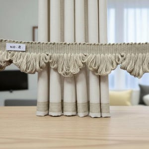 May include: A beige curtain featuring a horizontal trim with a looped fringe edge. A small white rectangular tag attached to the trim displays the text NO. 2. The curtain fabric has a vertical pleated design with a textured band near the bottom edge.