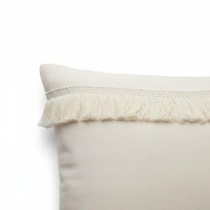 Op de afbeelding: Een crèmekleurig sierkussen met een gestructureerd stoffen oppervlak en een horizontale strook bijpassende franjes die over het bovenste deel van de kussenhoes is gestikt, wat een gelaagd visueel effect op het effen textiel creëert.