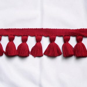 May include: Red tassel trim with a woven header. The trim features a series of evenly spaced, vibrant red tassels suspended from a matching red woven band. This decorative trim is suitable for various craft and sewing projects.