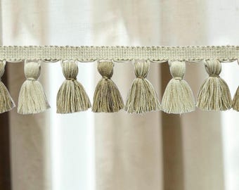 Peut inclure: Une bordure en tissu tissé beige présentant une bande supérieure horizontale avec une série de pompons suspendus dans des nuances variées de brun clair, de beige et de crème. Les pompons sont régulièrement espacés le long du bord inférieur.