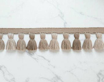 Peut inclure: Une garniture décorative avec une série de pompons beiges suspendus à une bande tissée. Les pompons varient en teinte, du beige clair au beige foncé, créant un effet de dégradé subtil. Idéal pour les projets de couture.
