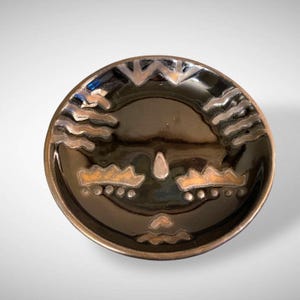 May include: A small black ceramic dish with a decorative design in gold. The design features a stylized face with wavy lines and dots.