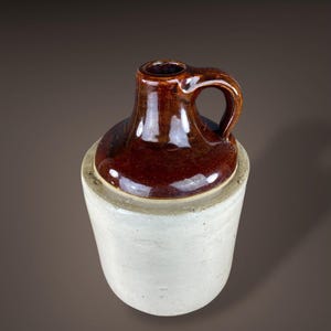 May include: A brown stoneware jug with a handle, set atop a cylindrical, off-white base. The jug has a glossy finish, and the handle is arched. The base is a lighter, matte color, creating a contrast. The jug is likely for storing liquids.