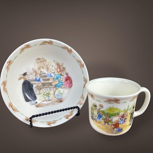 May include: A white ceramic bowl and mug set featuring illustrations of rabbits in various scenes. The bowl has a rim with rabbit silhouettes and a central image of rabbits. The mug has a handle and a similar rabbit scene. The set is displayed on a black metal stand.