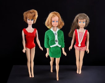 May include: Three vintage Barbie dolls wearing different outfits. The doll on the left is wearing a red swimsuit. The doll in the middle is wearing a green crocheted dress. The doll on the right is wearing a red and white striped swimsuit.