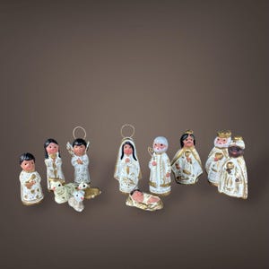 May include: A collection of hand-painted ceramic nativity figurines. The figures are primarily white with gold accents and depict various biblical characters, including angels, shepherds, and the three wise men. The figures are arranged on a brown background.
