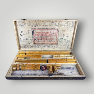 May include: An open wooden box containing vintage letterpress printing blocks. The box is aged with visible wear and tear, and the interior is divided into compartments. The lid features the text "ARISTOCRAT SIGN MARKER".