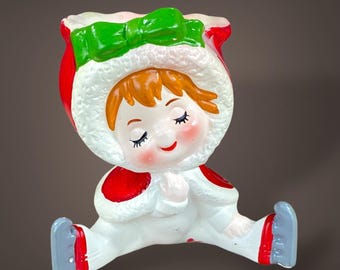 May include: A vintage Christmas figurine of a seated child in a white snowsuit with red accents. The child has orange hair, closed eyes, and a red smiling mouth. The hat is red with a green bow.