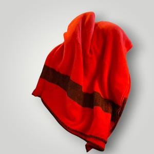 May include: A vibrant red blanket featuring a single horizontal dark brown stripe across the lower section. The fabric appears soft and folded, with a finished dark brown stitched edge along the bottom hem, creating a simple and clean design.