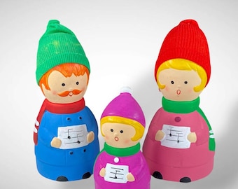 May include: Three ceramic carolers, each with a unique color scheme. The figures wear knitted hats, scarves, and coats in blue, pink, and purple. Each holds a sheet of music. The figures have painted faces with rosy cheeks and are a festive holiday decoration.