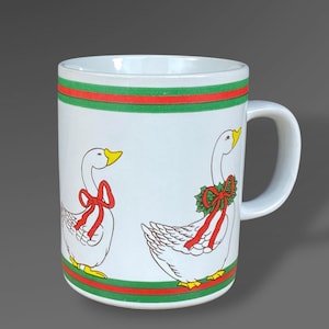 May include: White ceramic mug with a handle, featuring illustrations of geese with red bows and green accents. The mug has a red and green striped border. The geese have yellow beaks and feet. A decorative item.