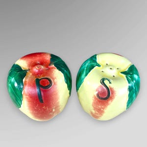 May include: A pair of ceramic salt and pepper shakers shaped like apples. One is red with a black "P", the other is yellow with a black "S". Both have green leaf accents.