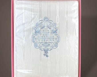 May include: A white photo album with a textured, striped cover encased in a pink box. The center features an intricate, light blue embossed design with floral motifs, birds, and the text "OUR WEDDING" written in a decorative, stylized font.