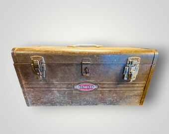 May include: A vintage Craftsman metal toolbox with a weathered, rusty appearance. The toolbox has a rectangular shape with a handle on top and two latches on the front. The Craftsman logo is visible on the front of the toolbox.