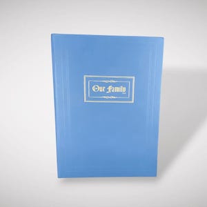 May include: A light blue photo album with the words "Our Family" in gold lettering. The album has a rectangular shape with a decorative border. The cover is a solid light blue color.