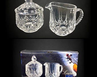 May include: A clear crystal sugar bowl with a lid and a matching creamer pitcher, both featuring a diamond-cut pattern. The set is displayed alongside its original blue cardboard box, which shows an image of a violin and the text Longchamp.