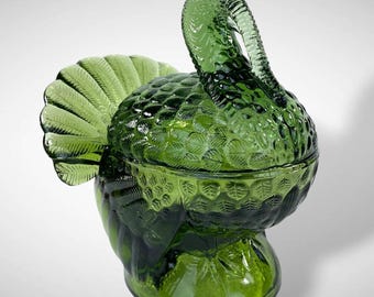 May include: A translucent forest green glass turkey-shaped covered dish. The piece features a detailed molded texture representing feathers on the body and tail, with a curved neck and head forming the lid handle, resting on a circular base.