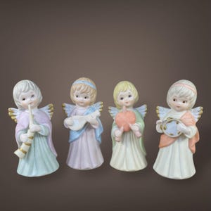 May include: Four ceramic angel figurines in pastel colors. Each angel has a different musical instrument: a flute, a lute, a fruit, and a tambourine. The angels have gold-colored wings and are wearing robes.