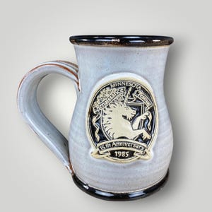 May include: A light gray ceramic mug with a dark brown rim and base. The front features a raised emblem of a unicorn with the text Minnesota Renaissance Festival 15th Anniversary 1985. The mug has a curved handle and a smooth, speckled glaze finish.