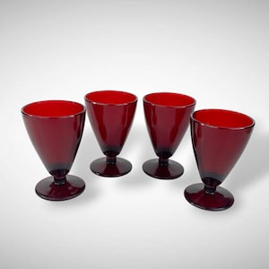 May include: Four ruby red stemmed glasses with a conical bowl and a circular base. The glasses are made of a dark red glass and are evenly spaced against a white background. The glasses are likely for serving beverages.
