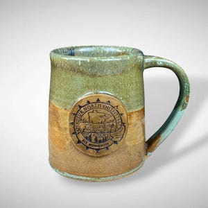 May include: A handcrafted ceramic mug with a unique, earthy color scheme. The mug features a green and brown glaze with a circular emblem that reads "Voyageur North Outfitters, Ely Minnesota". The handle and rim have a turquoise hue.