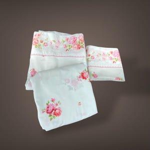 May include: White bed sheets with a floral pattern featuring pink and red roses. The sheets have a decorative border with a pink trim. The set includes a fitted sheet, a flat sheet, and a pillowcase.