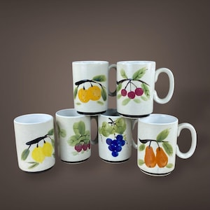 May include: A set of six ceramic mugs with fruit illustrations. Each mug features a different fruit design, including oranges, cherries, grapes, lemons, and pears, with green leaves. The mugs have a speckled beige background and black rims.