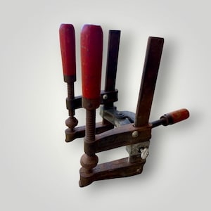May include: Three vintage woodworking clamps with red wooden handles. The clamps are made of metal and show signs of wear, with a rusty appearance. The handles are cylindrical and painted red. The clamps are arranged in a group, suggesting they are for holding materials together.