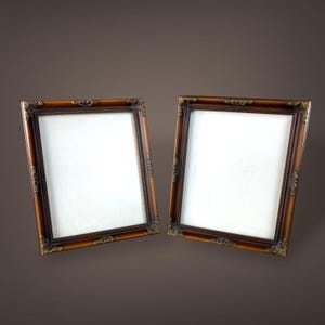May include: Two ornate, rectangular picture frames with a dark brown finish and gold-colored corner accents. The frames have a classic, vintage design, with a white interior space for displaying photos or artwork. The frames are set against a brown background.