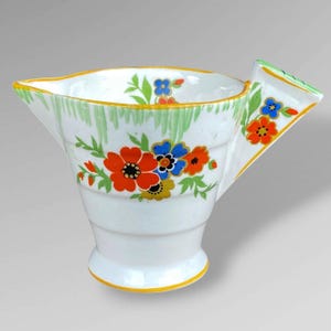 May include: A white ceramic creamer featuring a hand-painted floral pattern with vibrant red, blue, and yellow flowers, green leaves, and a yellow rim. The vessel has a wide spout, a flared base, and a distinct angular handle with green accents.