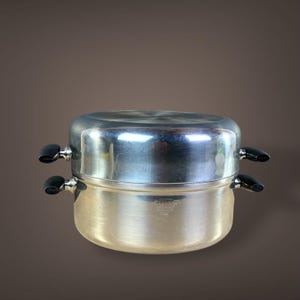 May include: A two-tiered stainless steel steamer pot with black handles. The pot has a shiny, reflective surface and is designed for cooking various foods. The handles are on the sides of the pot, and the pot is sitting on a brown background.