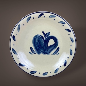 May include: A white ceramic plate with a blue apple design in the center. The plate has a blue border and is decorated with blue teardrop shapes around the rim. The plate is round.