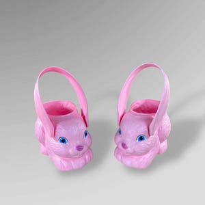 May include: Two pink Easter bunny-shaped baskets with handles. The baskets have blue eyes and a pink nose. The baskets are made of plastic and are designed to hold Easter eggs or treats.