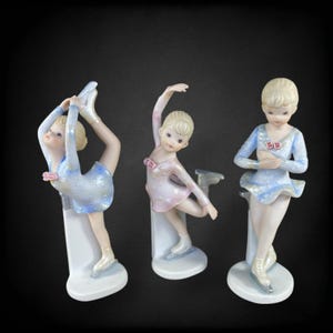 May include: Three porcelain figurines of ice skaters in graceful poses. Each figure wears a light blue or pink dress with silver accents and ice skates. The figurines are on white bases against a black background.