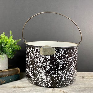 May include: A vintage black and white speckled enamelware bucket with a metal handle. The bucket has a white interior and a metal clasp. The handle is arched and made of metal. The bucket is a classic kitchenware item.