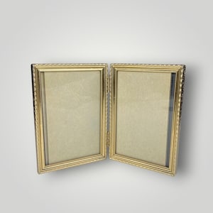 May include: A gold-colored, hinged double picture frame. The frame has a decorative border and is open, revealing the interior space for photos. The frame is set against a plain, light background.