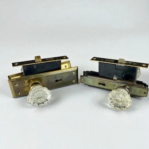 May include: Two vintage door lock sets. Each set includes a brass plate, a black lock mechanism, and a clear, flower-shaped glass doorknob. The hardware is designed for installation on a door and is likely from an older home.