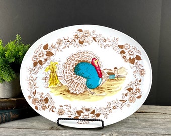 May include: White oval serving platter with a brown floral border and a colorful illustration of a turkey in the center. The turkey is blue, brown, and red with a yellow beak and red wattle. The background of the illustration is a yellow field with a brown barn in the distance.
