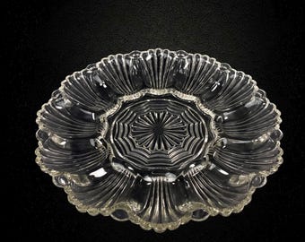 May include: A clear glass serving platter featuring a central circular medallion with radiating lines, surrounded by a ring of twelve distinct, shell-shaped compartments with scalloped edges, creating a decorative and functional dish.
