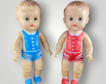 May include: Two vintage plastic dolls with molded hair and painted facial features. One doll wears a blue one-piece outfit with white buttons, while the other wears a red one-piece outfit with white buttons. Both dolls have matching colored shoes.