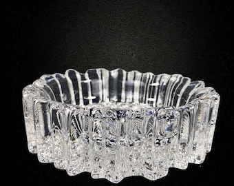 May include: A clear glass bowl featuring a thick, heavy base and a series of vertical, textured ridges along the exterior wall. The circular vessel has a scalloped, wavy rim and a transparent, reflective surface that catches light across its facets.