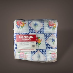 May include: A Cannon Monticello Floral Patch blanket, wrapped in clear plastic. The blanket features a patchwork design with floral patterns in red, yellow, and blue, set against a white background with blue geometric borders. The label is red and white.