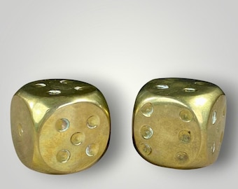 May include: Two solid brass dice with rounded corners and polished surfaces. The left die shows five circular pips arranged in a quincunx pattern on its front face, while the right die displays six circular pips in two vertical rows.