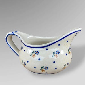 May include: Cream-colored ceramic creamer with a blue rim and handle. The creamer is decorated with blue polka dots and small floral clusters in blue and orange. It has a curved spout for easy pouring.