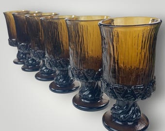 May include: A set of six amber-colored glass goblets arranged in a diagonal line. Each goblet features a textured, vertical bark-like pattern on the bowl, a decorative band of raised geometric shapes, and a thick, twisted stem on a circular base.