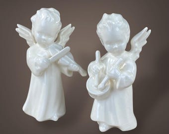 May include: Two white ceramic angel figurines. One angel plays a violin, the other plays a drum. Both angels have wings and are dressed in robes. The figurines are set against a brown background.