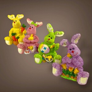 May include: Four colorful Easter bunny figurines. The bunnies are yellow, pink, green, and purple. Each bunny holds an Easter-themed item, such as carrots, a duckling, an Easter egg, and flowers. The figurines are sitting down.