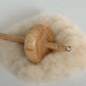 May include: A wooden drop spindle with a light-colored, rounded whorl and a slender shaft, resting on a cloud of soft, undyed wool fibers. The spindle is designed for hand spinning yarn, a traditional craft.